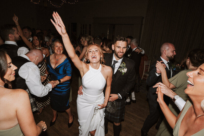 bride holding phone to take a selfie of her and groom, sticking toungues out bride wears black hate and groom wears red blazer and sunglasses
