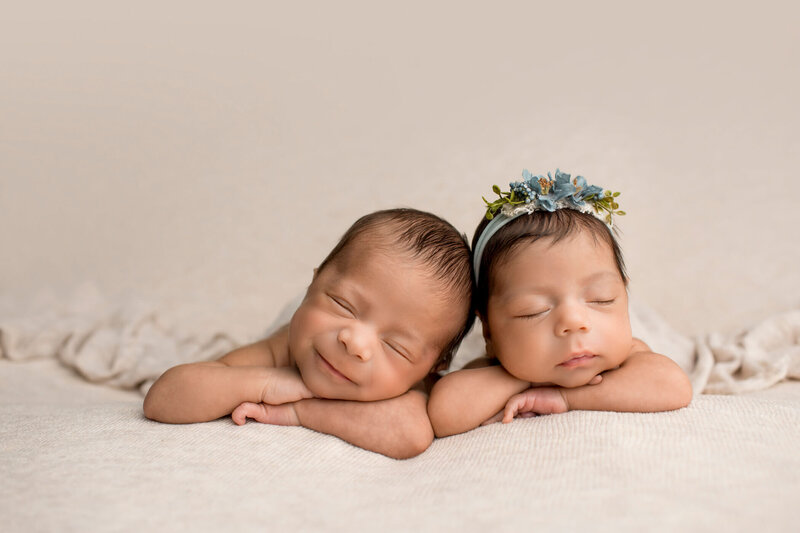 A newborn baby is peacefully sleeping, curled up on a soft, dusty rose-colored background. The baby is naked, showcasing their delicate, natural form with adorable baby rolls. A floral headband with pink and green flowers adorns the baby's head, adding a touch of sweetness to the serene scene. The baby's tiny hands are gently placed near their face, and their expression is calm and content, creating a heartwarming and tender portrait.