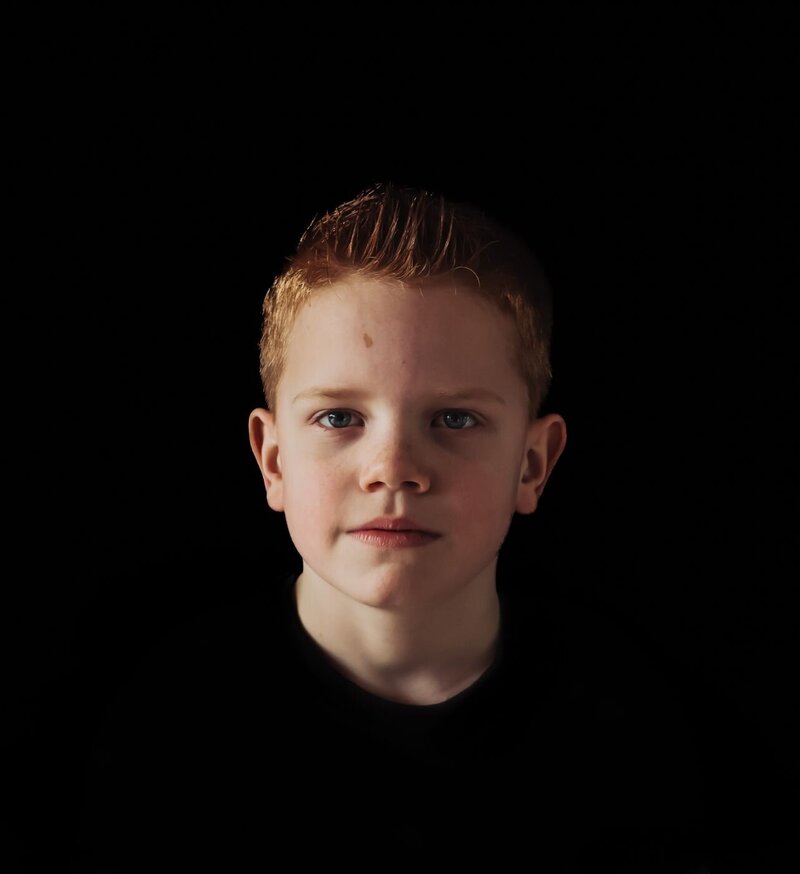 Een portretfoto van een jonge jongen met een open en vriendelijke uitstraling, gefotografeerd tegen een donkere achtergrond