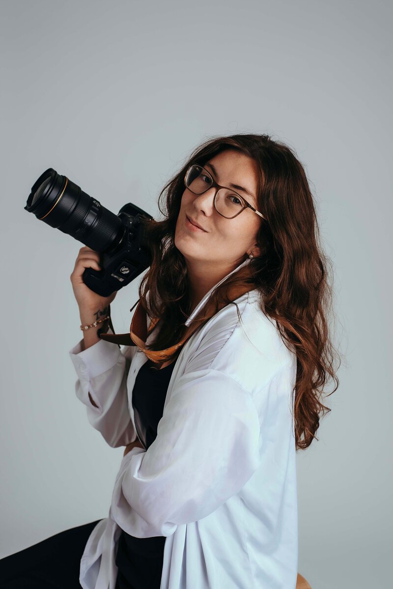 A branding photo of Mobile, AL Family photographer. She is looking at the camera with confidence, as she holds her camera in her hand. She is wearing a black shirt under a white button up.