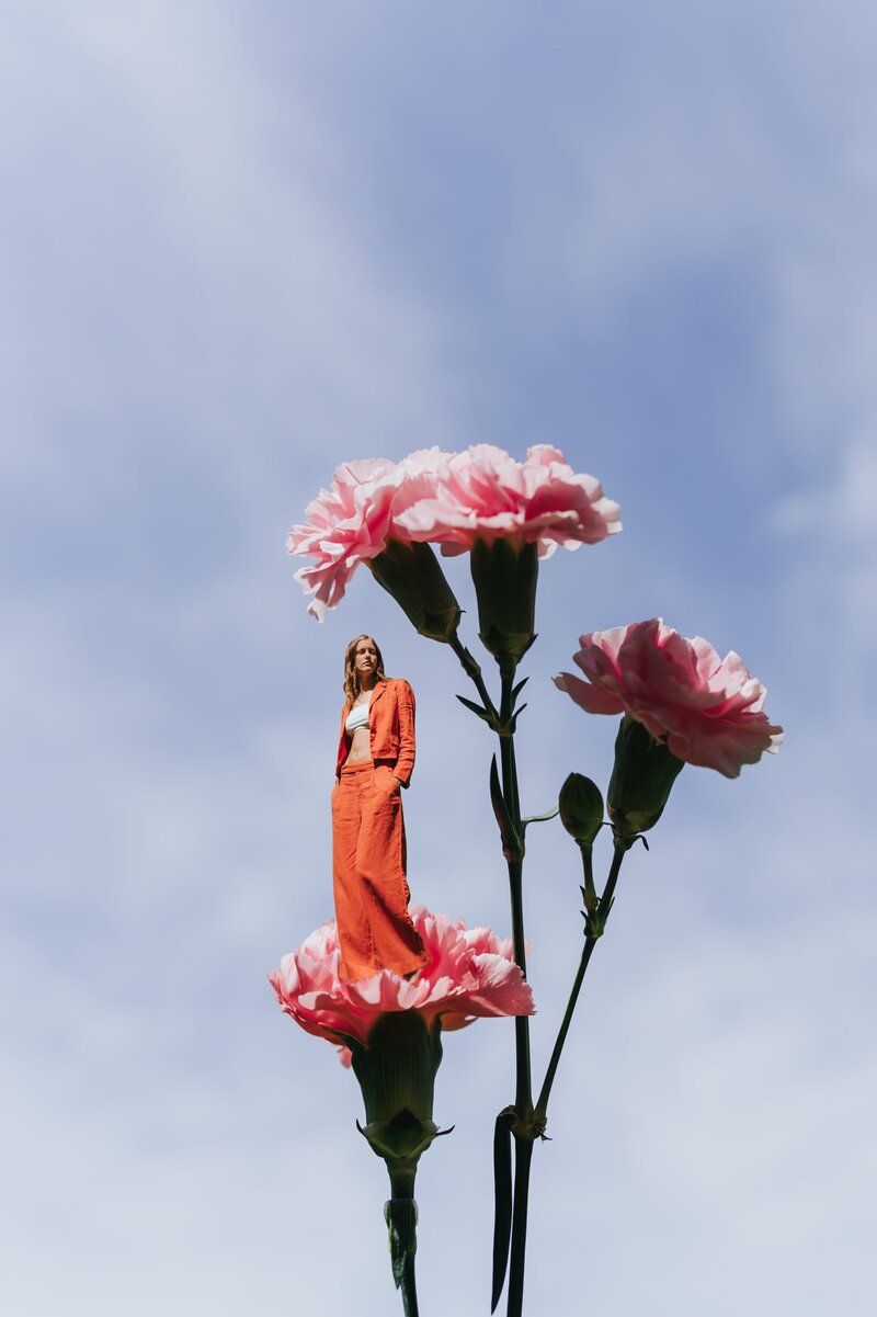 Woman in orange suit is photoshopped to seemingly stand on top of a pink carnation flower with blue skies in the background