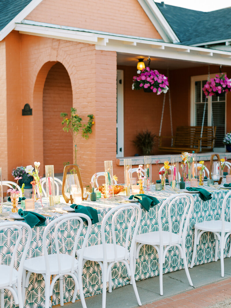 white floral arrangement with hot pink candle sticks and teal napkins on a table with a pink and teal printed tablecloth in front of an orange historic house