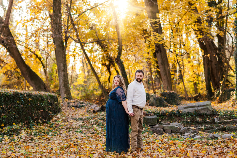 A pregnant woman standing back to back with her partner in a wooded area.