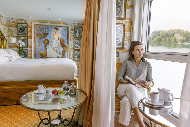 A woman sits by a large window in a bright, elegantly decorated cruise ship cabin, reading a book. A bed, artwork, and a glass table with fruit and tea are visible in the room, with water and greenery outside the window.