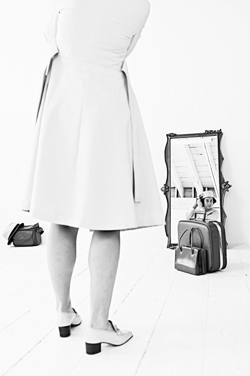 A black and white photo of a woman's reflection in a mirror with luggage in front of it.