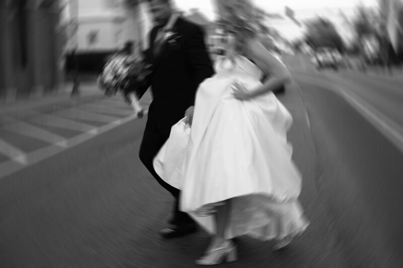bride and groom running across city street laughing while bride holds wedding dress up