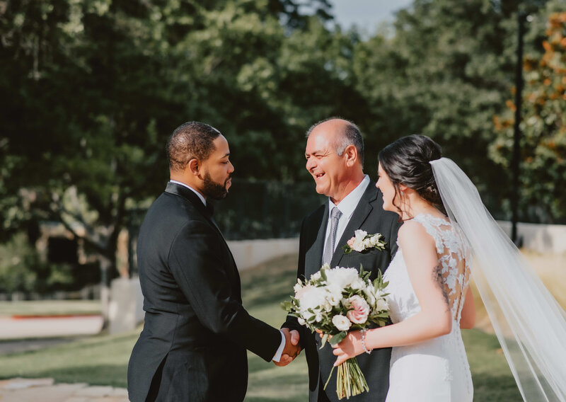 Outdoor wedding moment with a groom shaking hands with the bride’s father while the bride in a white gown and veil holds a bouquet of flowers, captured in natural sunlight.