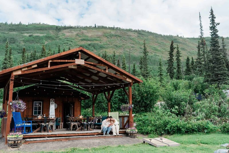 a couple sits on the edge of an outdoor deck area with a mountain in the background. They are in casual clothes on day two of their Alaska elopement, snuggled into each other, smiling.
