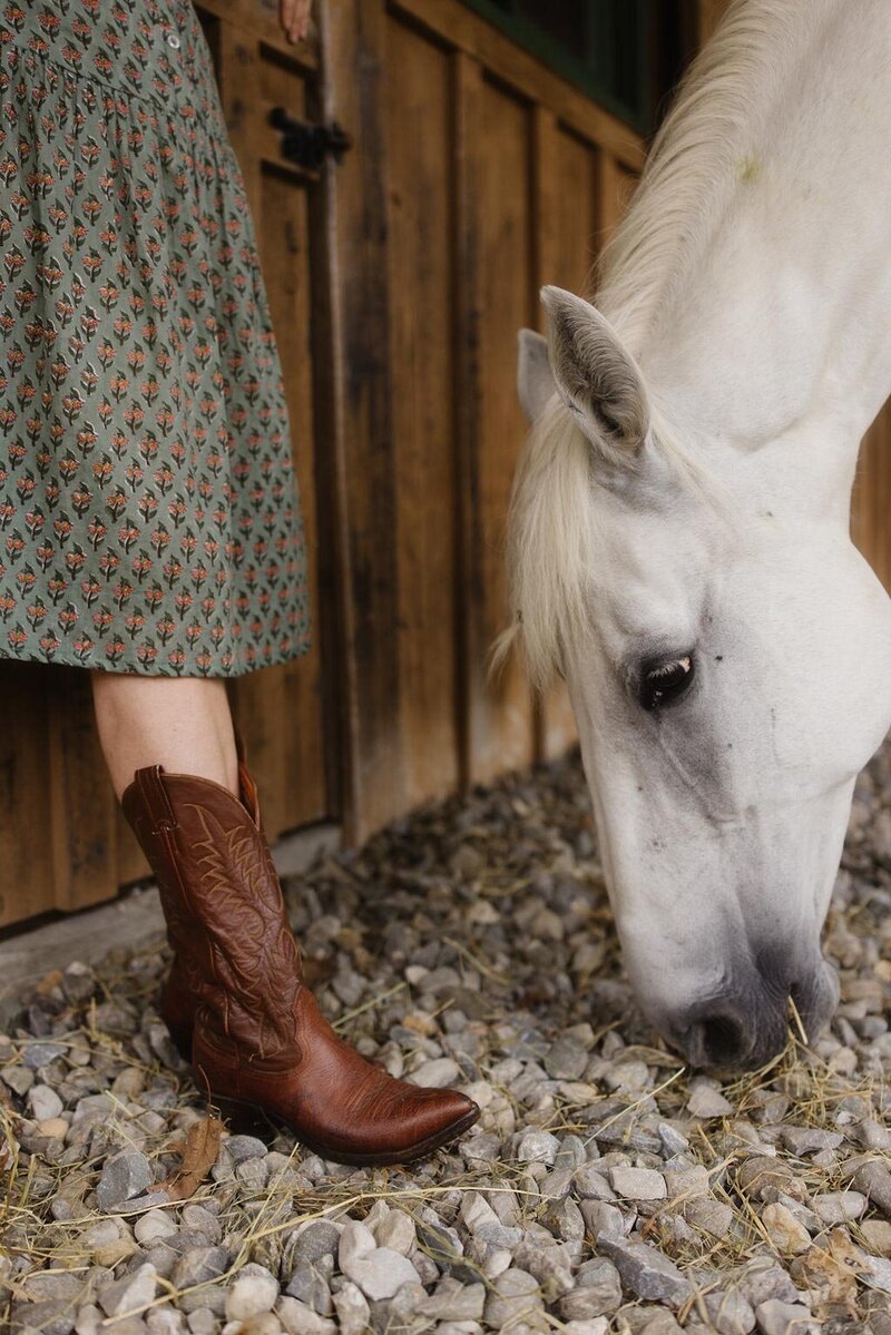 Detail of a white horse beside a woman in western boots, standing on stone outside a barn — symbolizing grounded connection and partnership in equine-assisted healing in the Berkshires