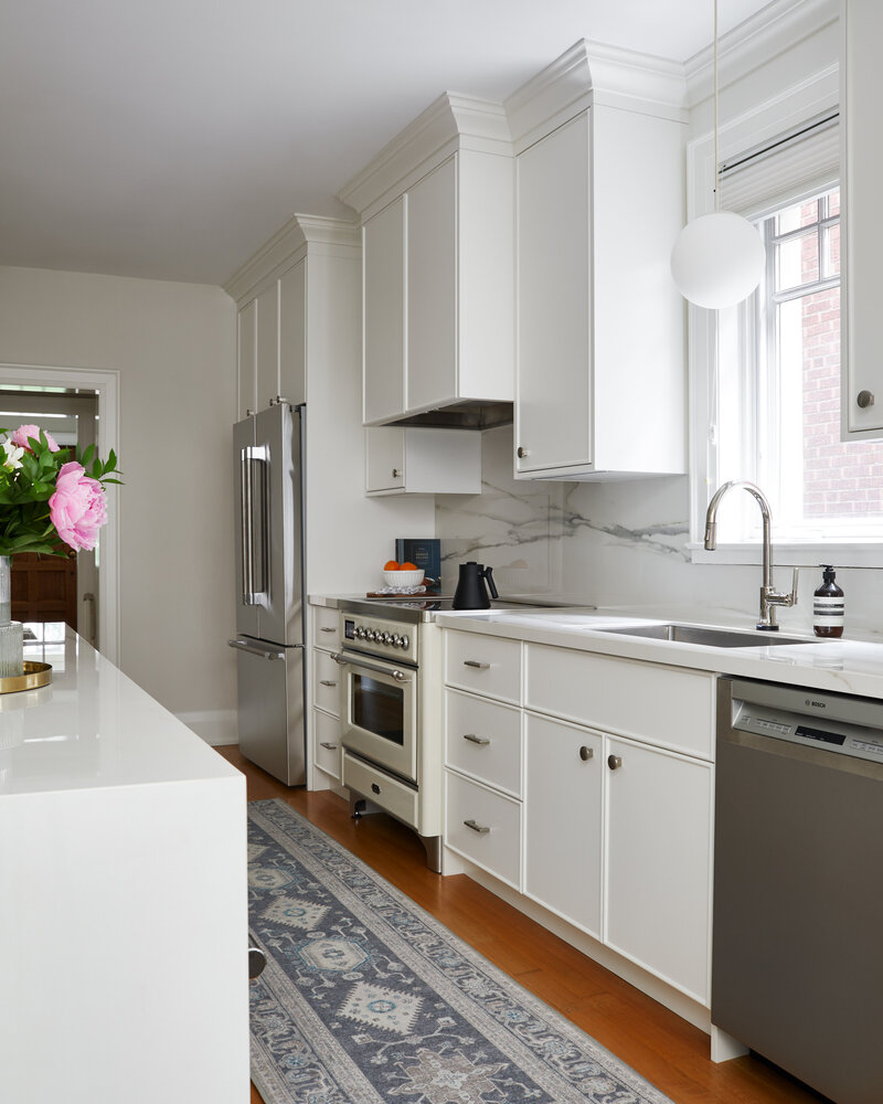 A bright airy kitchen with modern white cabinetry, wood flooring covered by a grey and white patterned rug, stainless steel appliances, and a globe pendant lighting fixture hanging from the ceiling