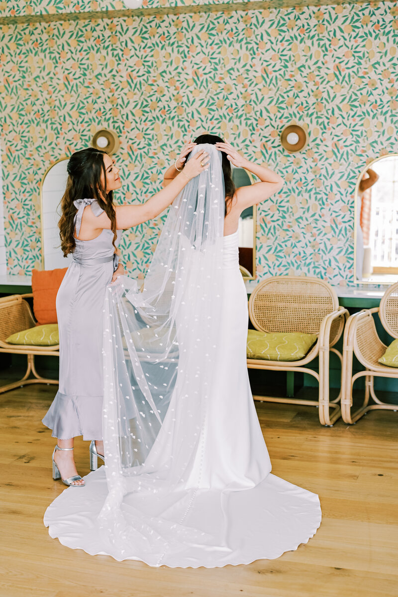 Bridesmaid helping bride put on her wedding veil in front of a wall with colorful wallpaper