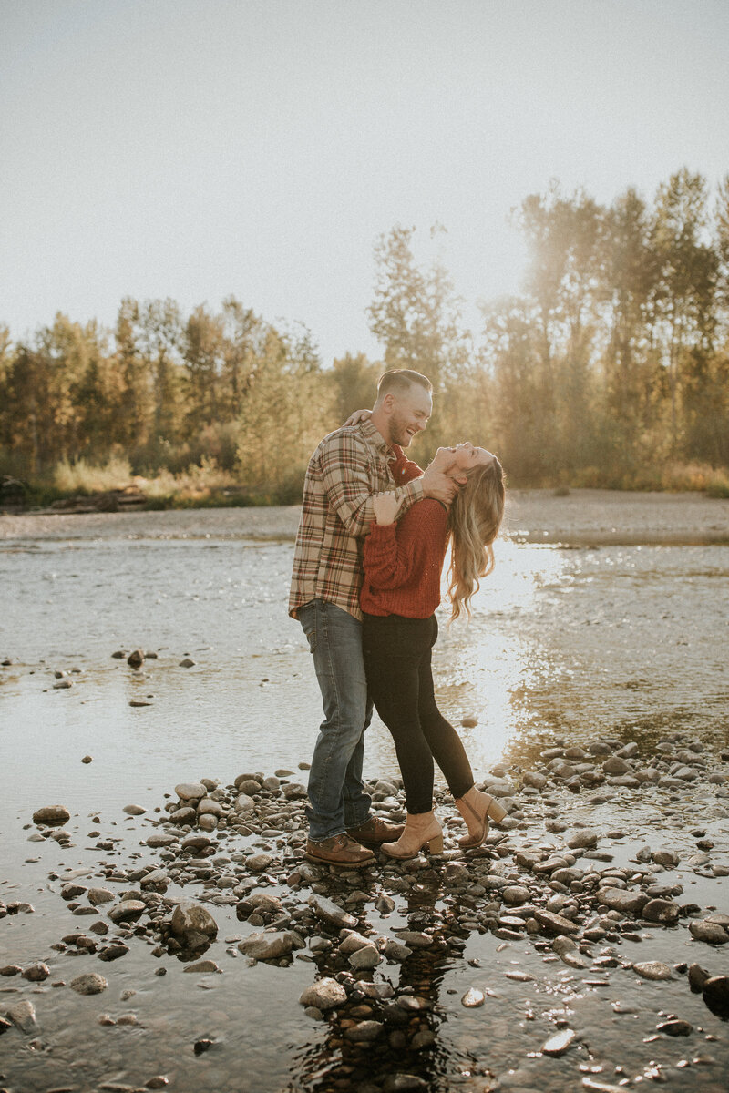 A man wraps his hand around his fiance's neck as she looks up laughing in front of a lake