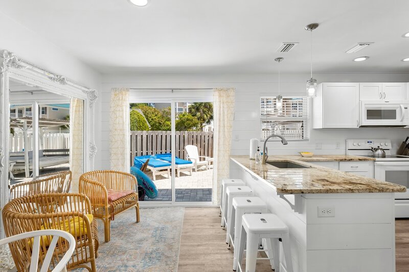 Coastal kitchen with granite countertops and island seating, overlooking a sunny patio with lounge chairs and private fenced yard.