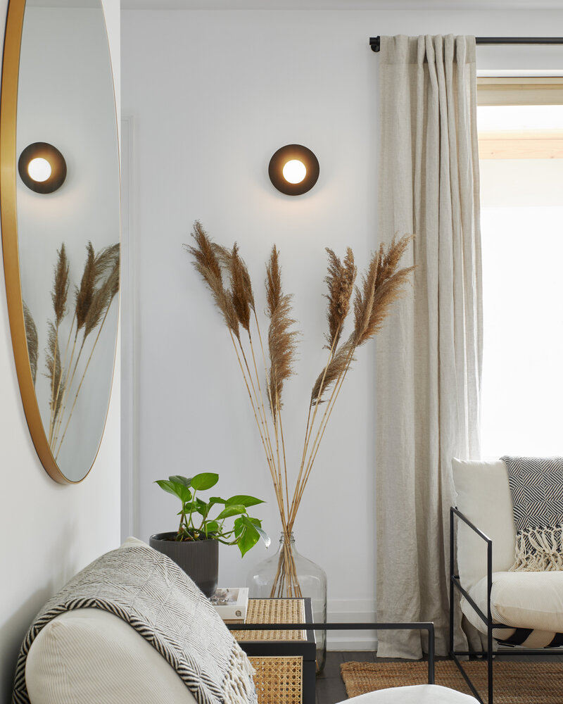 A bright living room area with earthy tones featuring a wall-mounted globe pendant light with a dark base, tall dried plants in a glass bottle, a seating area with black and white patterned throw blankets and chairs, a potted plant, wall-mounted mirror, and a tall window with flowy beige curtains.