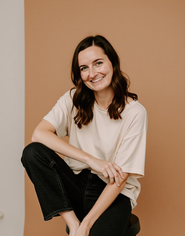 A woman named Jordin Brinn, a web designer, sits on a stool wearing jeans and a off-white shirt.