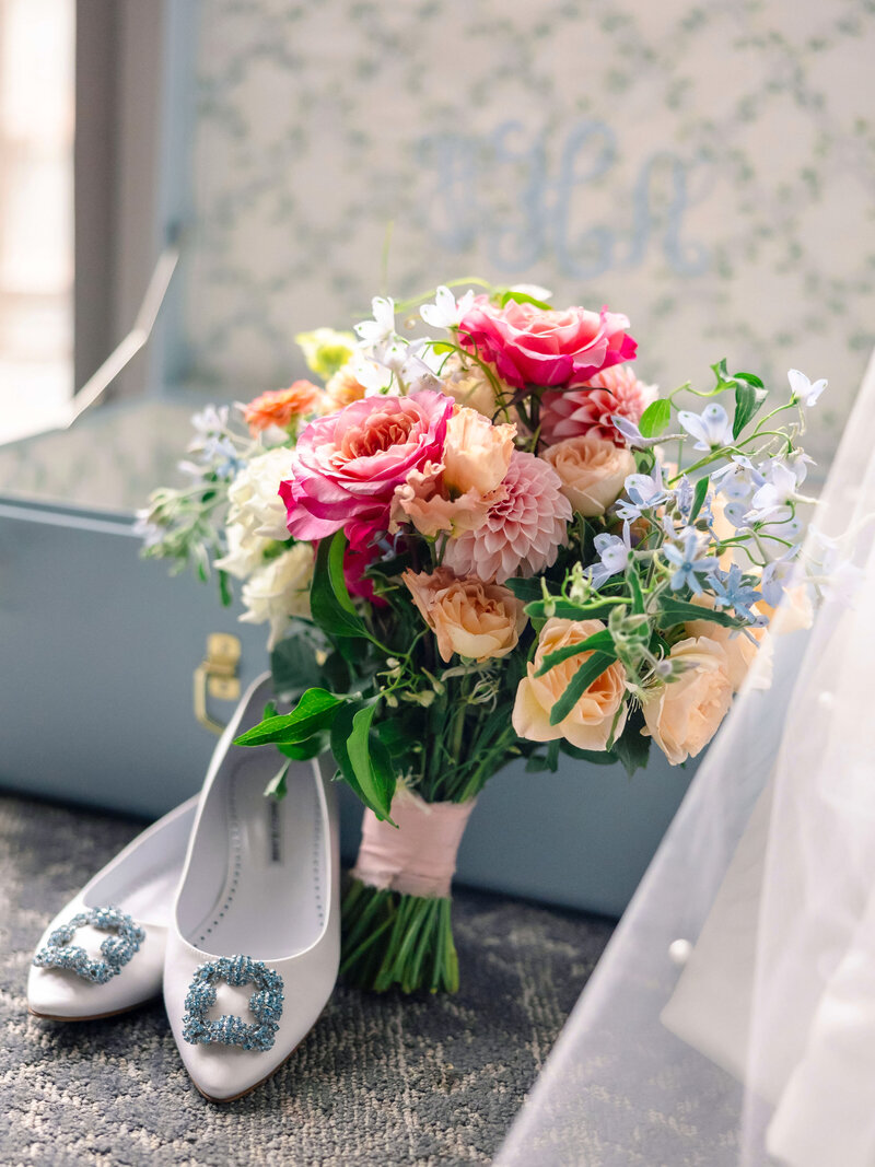 A bouquet of colorful flowers, including pink roses and light blue blossoms, is displayed next to a pair of white high-heeled shoes with jeweled embellishments. The background shows a light blue box with a subtle floral pattern.