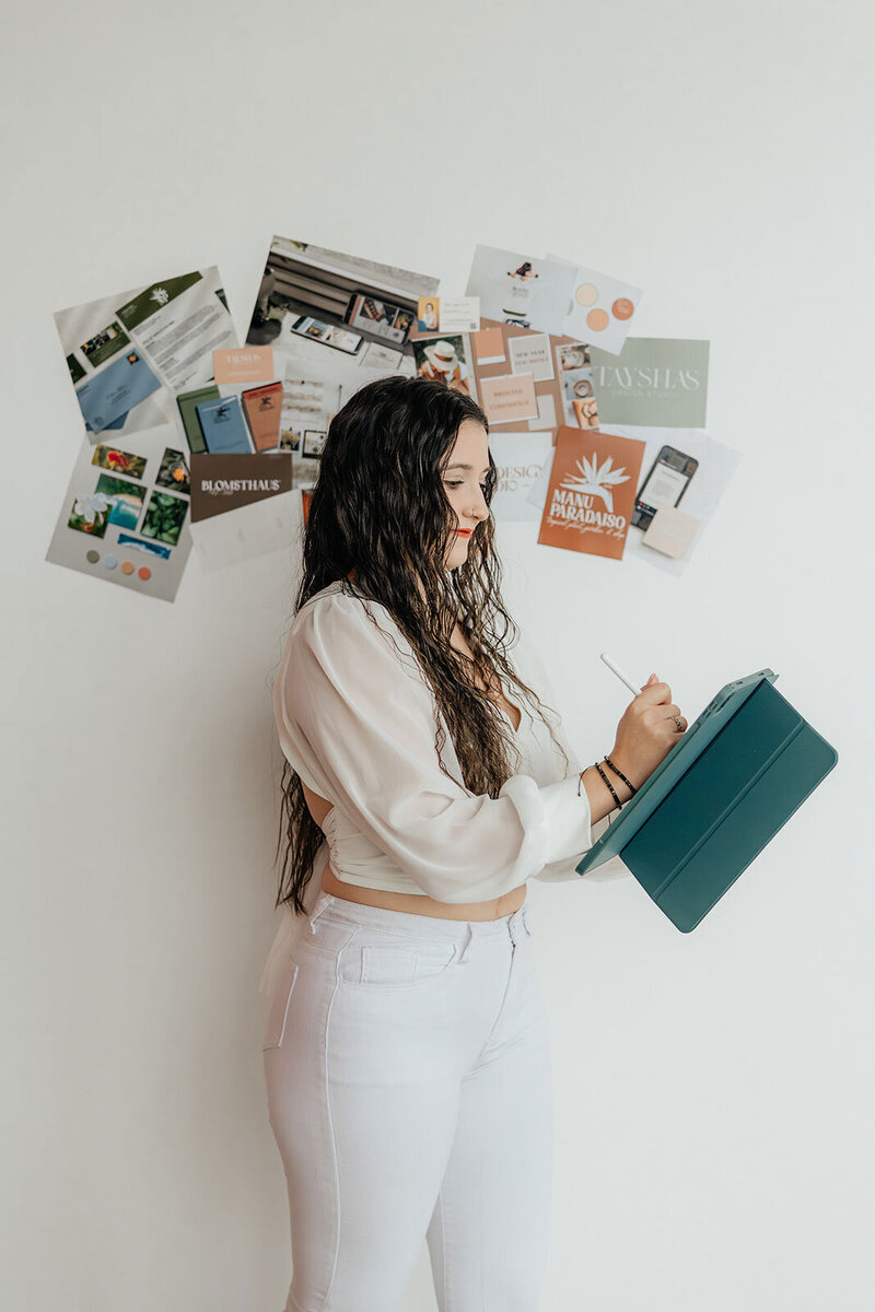 Taysha working on her tablet leaning against a white wall with a brand moodboard hanging behind her.