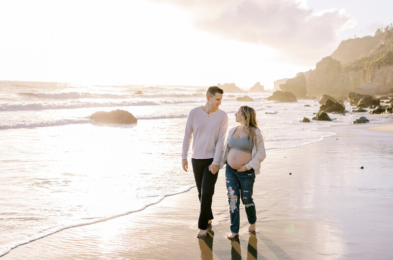 A couple holding hands and walking along the beach during their maternity portraits