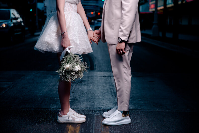 Bride in a short white dress holding a bouquet and groom in a light suit holding hands on a city street at night, photographed by Legacy in Light Photography.