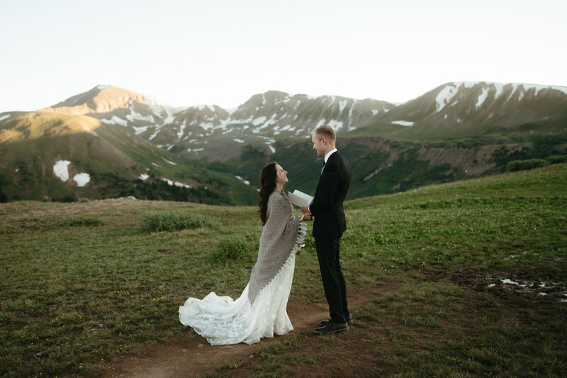 Hiking wedding ceremony, groom reads his vows to his bride with snow-covered mountains behind them