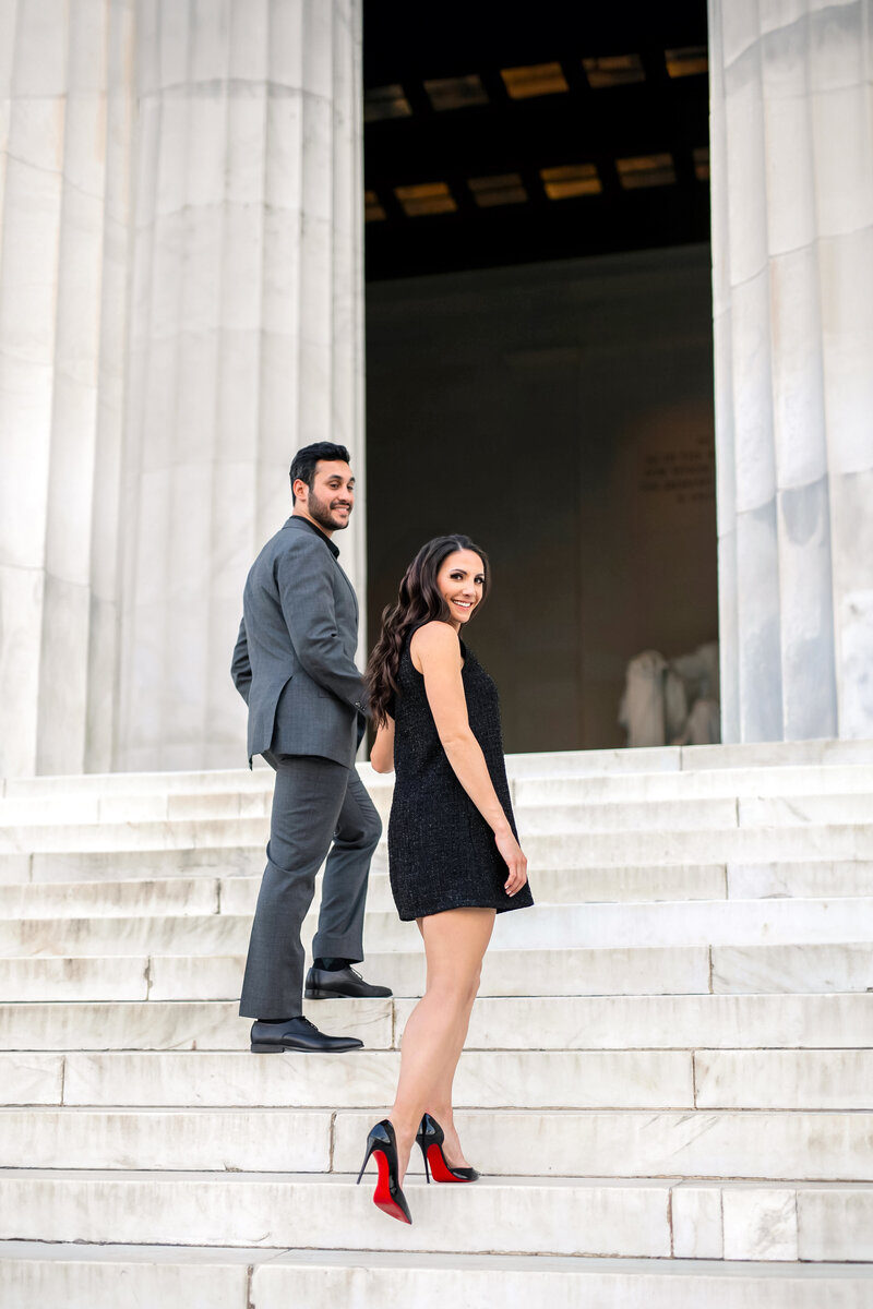 A man and woman in formal attire ascend marble steps outside a grand building with tall columns. The woman, in a black dress and heels, glances back, while the man, in a gray suit, leads the way.