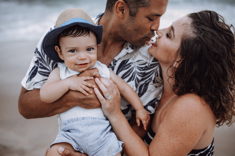 A mom and dad holding their young son lean in for a kiss at the beach