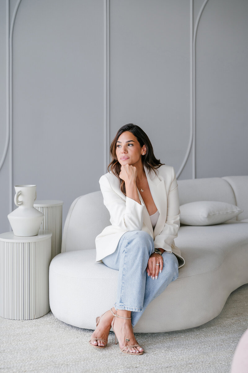 Valerie Roberson, interior designer at Interiors by CV, seated on a sofa with her hand resting on her chin, dressed in a white blazer and jeans, beside a modern side table.