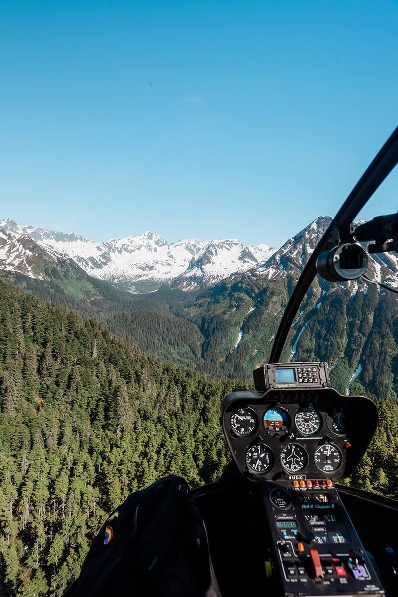 Rugged snow capped mountains are viewable out of the cockpit during private helicopter flightseeing tour headed to a private dogsledding experience for a luxury elopement in near Bear Glacier and Bear Lake in Seward.  There are swaths of rich green trees the valley between the rugged peaks, and the instruments of the helicopter are viewable in the lower right of the frame.