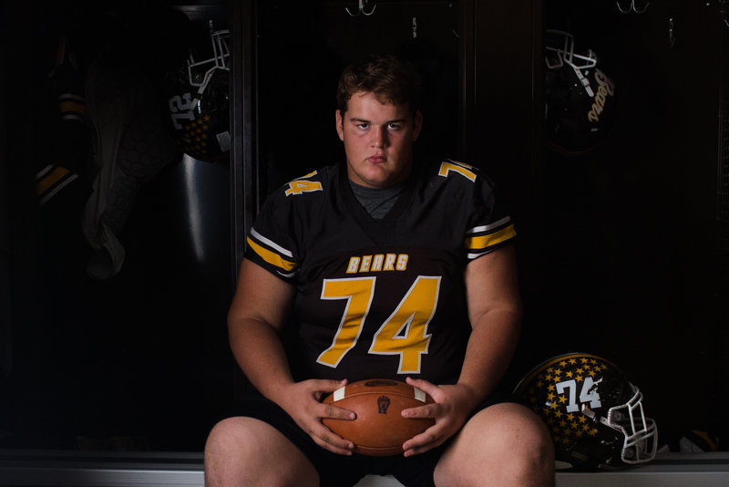High school senior guy in locker room, sitting on bench holding football wearing jersey and shorts, dimly lit room with dramatic lighting, photographed by Jamie Lynette Photography