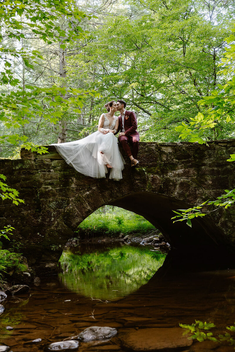 A couple kisses one another while sitting on a stone bridge over a river in the forest.