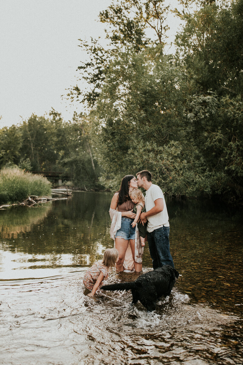 Husband and wife kiss as kids and dog splash around in a lake