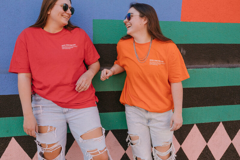 Two friends laughing while leaning up against a wall.
