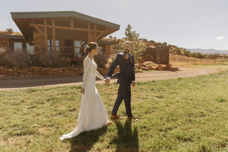 A groom holding the hand of his bride and leading her away from the camera as the walk together. Behind them, a small lodge made of tan, orange stone bricks.