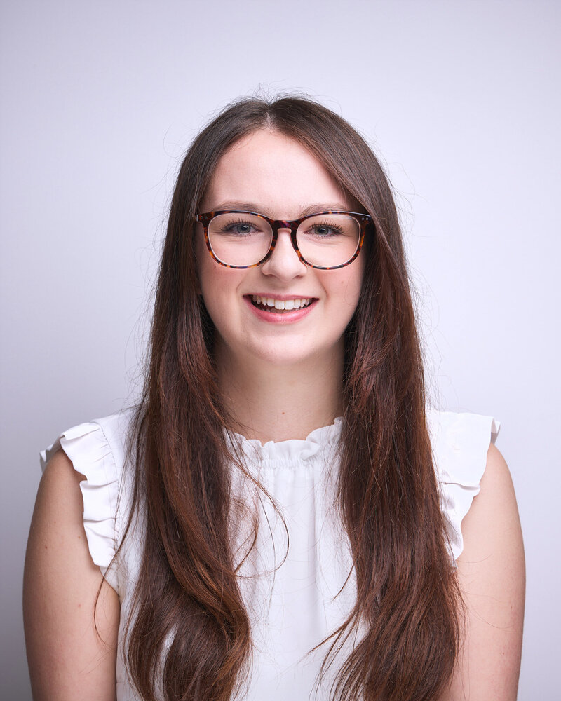 Woman with long hair, wearing glasses and smiling. She is indoors and the photo focuses on her face and upper body.