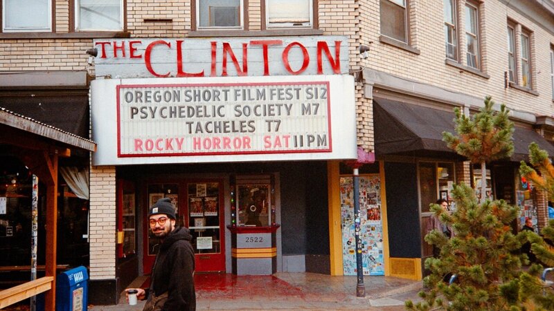 A man holding a coffee cup stands in front of the Clinton Theater, with a marquee listing events including 'Oregon Short Film Fest' and 'Rocky Horror' showing Saturday at 11 PM. The theater is housed in a brick building with posters and colorful decorations around the entrance.