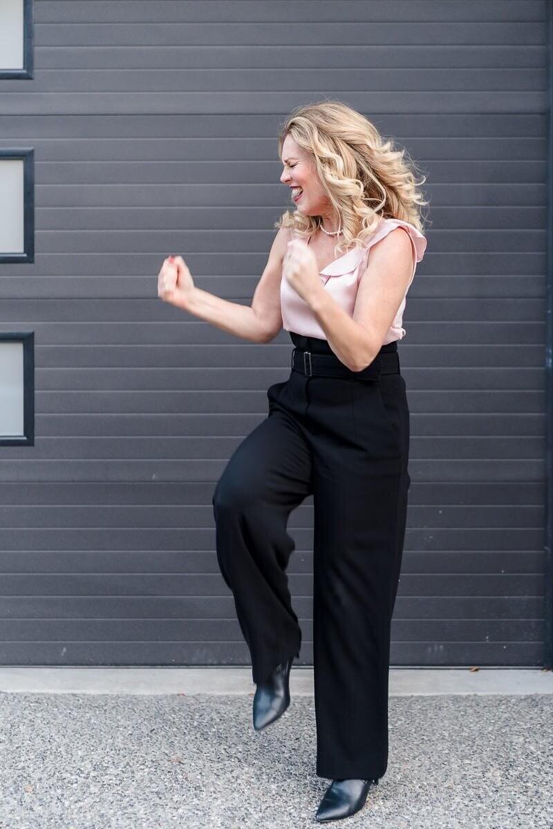 Professional woman in pink blouse and black trousers dancing excitedly in front of grey background