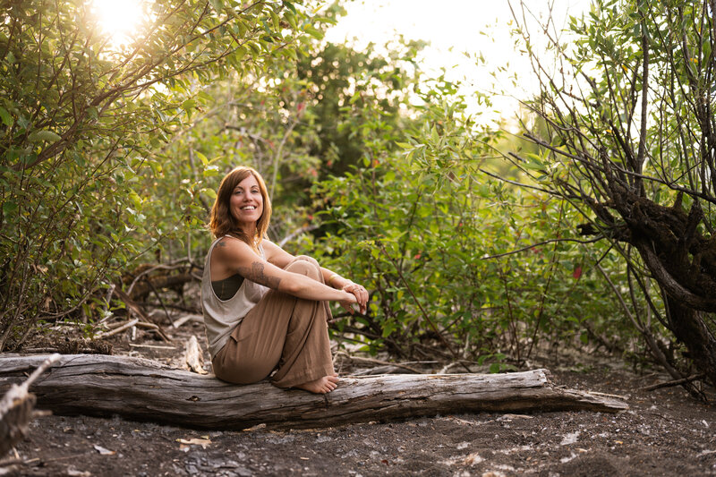 yoga teacher smiling at the camera from a log with green trees behind her