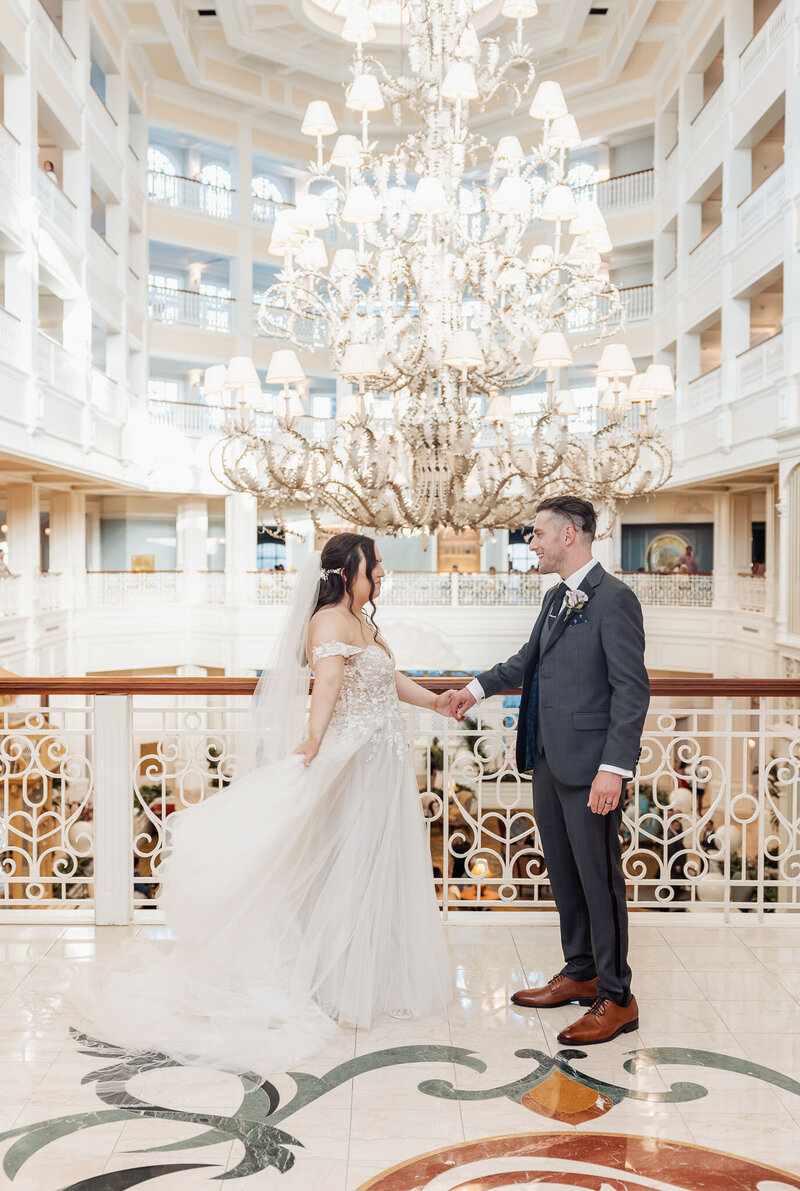 bride in a white gown with white veil and brunette hair stands on the left. She is holding hands with a man in a gray suit while he stands to the right. There is a large gold chandelier past the couple.