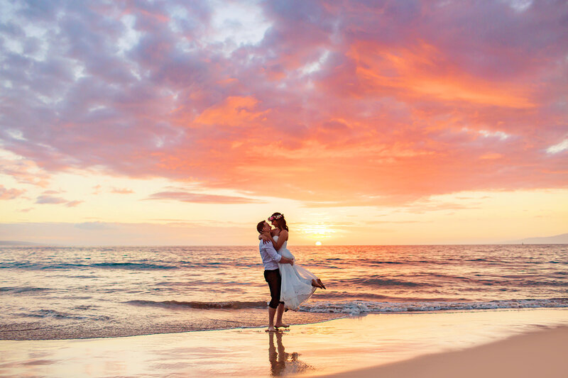 A couple embraces on a sandy beach in Maui at sunset, the sky glowing in soft pinks and oranges