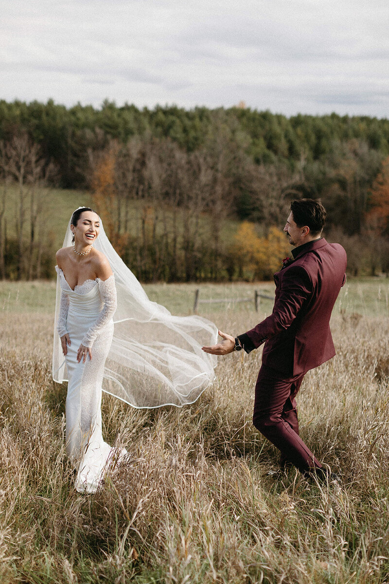 A bride in a white gown and veil laughs with a groom in a burgundy suit as they pose for an Ontario destination wedding photographer at Farmhill Weddings and Events in Peterborough, standing in a grassy field with trees.