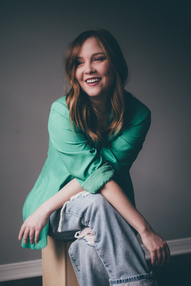 A smiling woman with long hair wears a green shirt and ripped jeans while sitting on a stool against a plain background, embodying the approachable vibe that website designers for small business aim to capture.