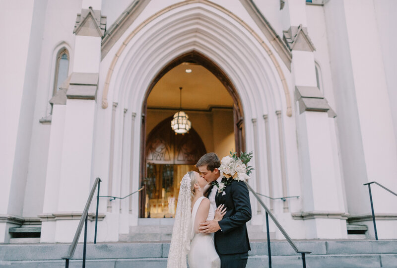 Bride and groom kiss in savannah wedding photograph in front of the cathedral basilica of st. john the baptist after recessional from wedding ceremony. Bride is wearing a spanish lace veil draping down her back and has florals of magnolia leaves and peonies cupped around the back of the grooms neck who is in a black tux.
