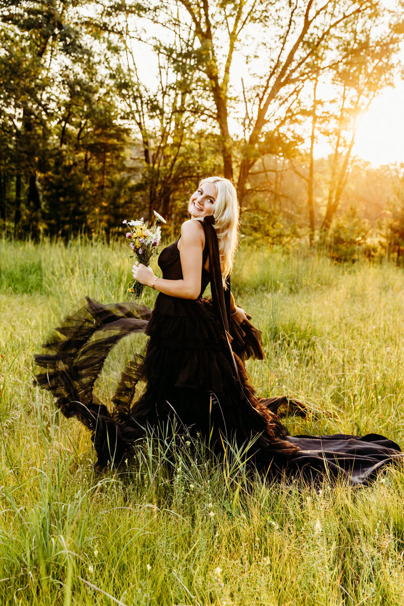 gorgeous teenage girl in black gown playing with her skirt as she holds a flower bouquet in a field near Green Bay