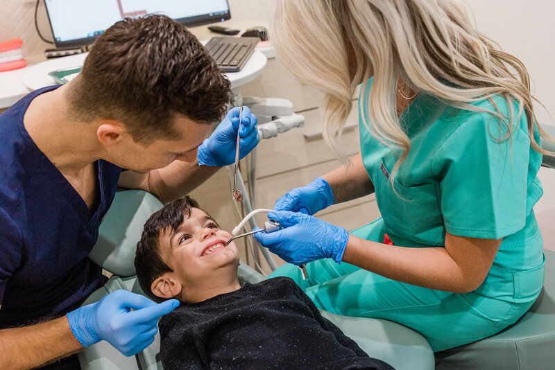 In his Andersonville dentist office, Dr. Michael Rabinowitz examines the teeth of a smiling young patient who is wearing a black heather t-shirt. Dr. Michael's dental hygienist assists with a water rinse and suction.