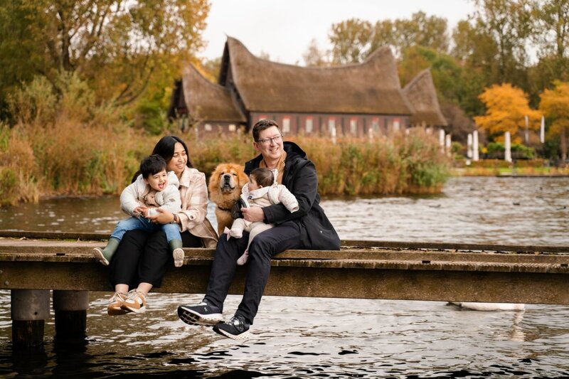 Mother holding hands with her young son while walking on a wooden pier beside swans and city skyline.