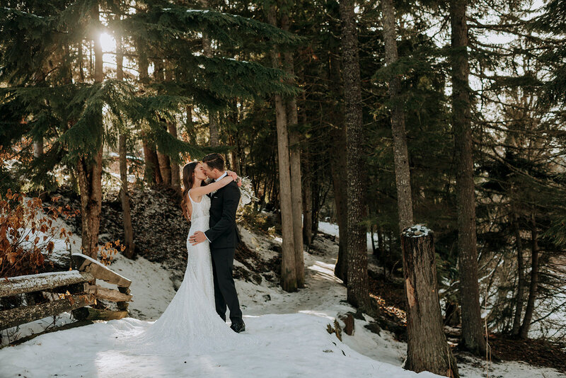 A bride and groom kiss on a walking trail with trees in the background and sun peaking through. The path is covered in snow.