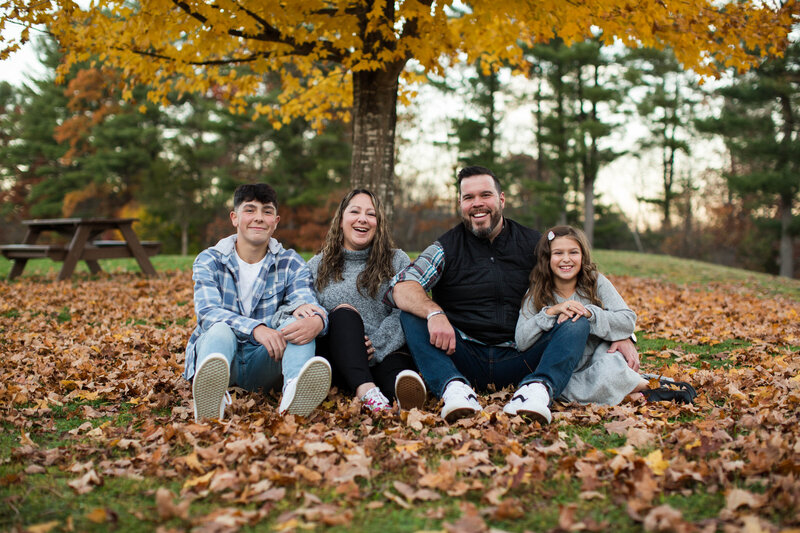family of four sitting in under the tree in the leaves in the fall