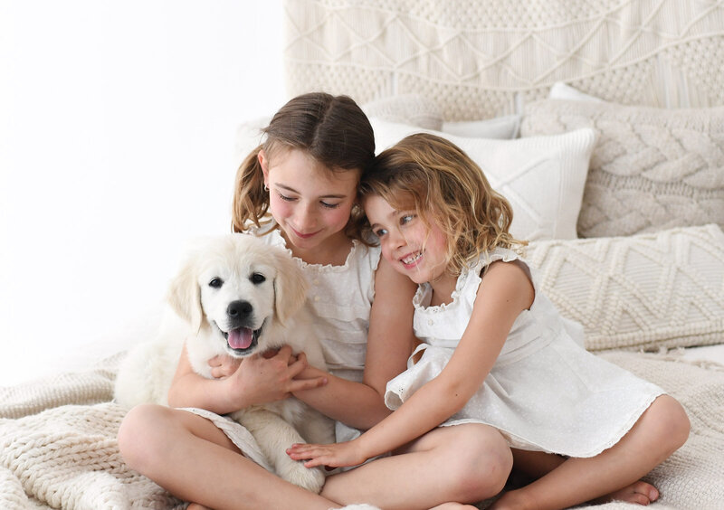 Two young girls sitting on a bed, both wearing white dresses. The older girl holds a fluffy white puppy that is panting and looking at the camera, while the younger girl smiles at the puppy. Perfectly captured by a St. Louis photographer, the background includes textured pillows and blankets in neutral tones.