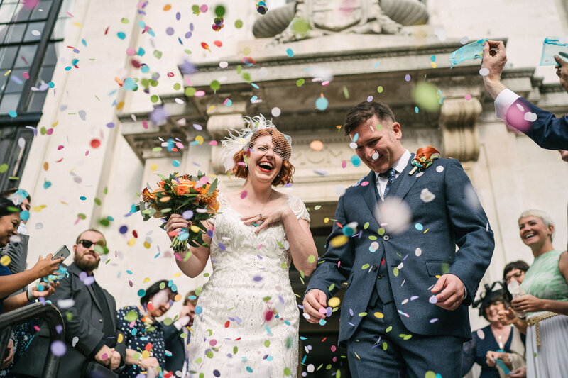 A colourful confetti moment with a bride and groom outside a town hall.