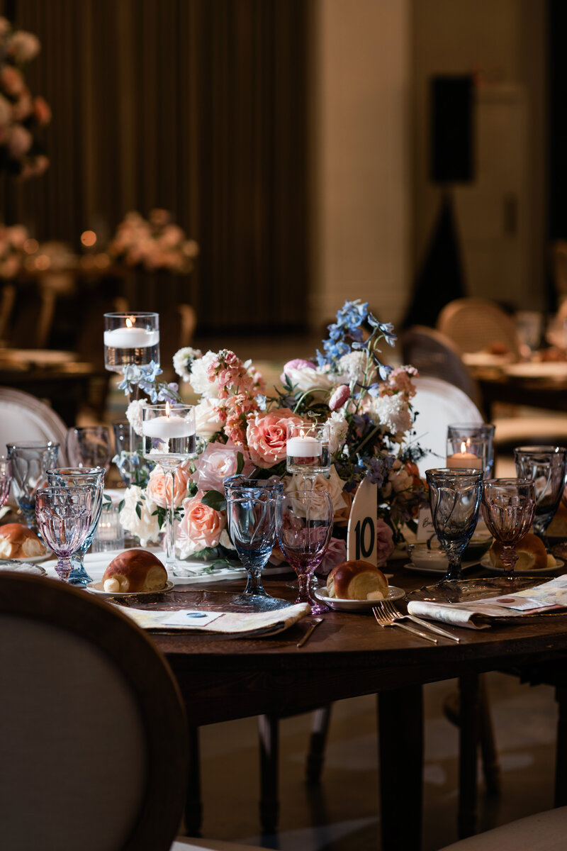 A round wooden farmhouse table decorated for a formal wedding with blue glassware, floral centerpieces, and place settings.